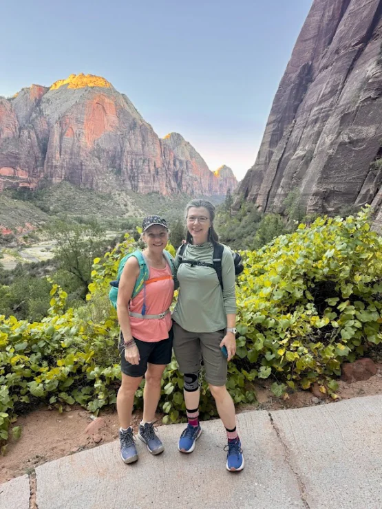 Two people stand on a trail with rocky cliffs and greenery in the background, both wearing hiking gear and smiling at the camera.