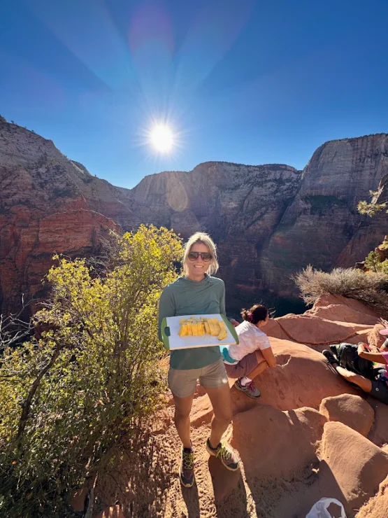 Person standing on a rocky ledge holding a plate of food, with a sunlit canyon in the background.