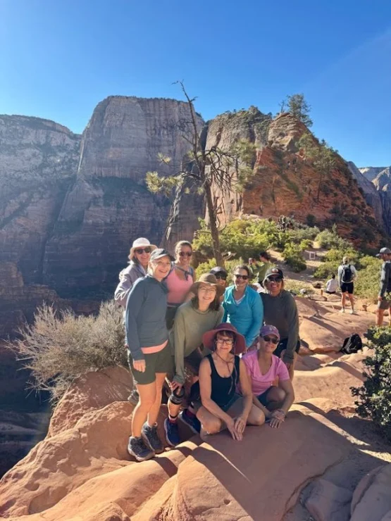 A group of seven people pose on a rocky ledge with a backdrop of cliffs and clear blue sky. They are wearing casual hiking attire.