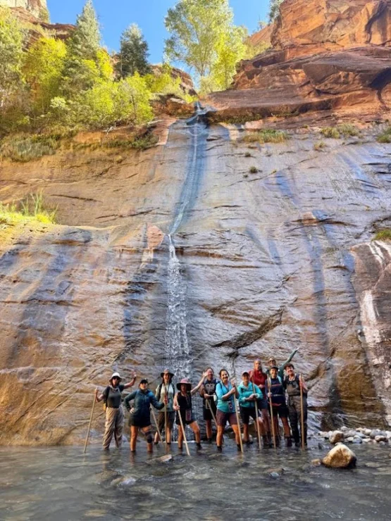 A group of hikers stands in shallow water at the base of a rock cliff with a narrow waterfall, under a clear blue sky.