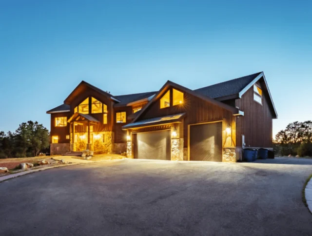 Large wooden house with illuminated windows at dusk, featuring two garages and surrounded by trees.